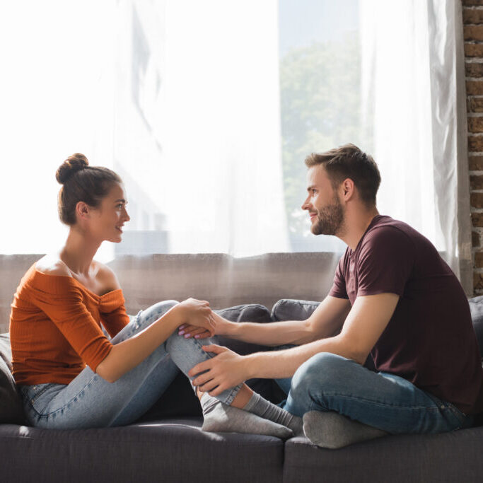 side view of young couple talking while sitting on sofa and holding hands